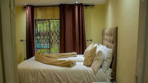 A bedroom with a bed, pillows, a bedside table with a lamp, and a window with curtains. - Chishongo Lodge for one (Darwendale)