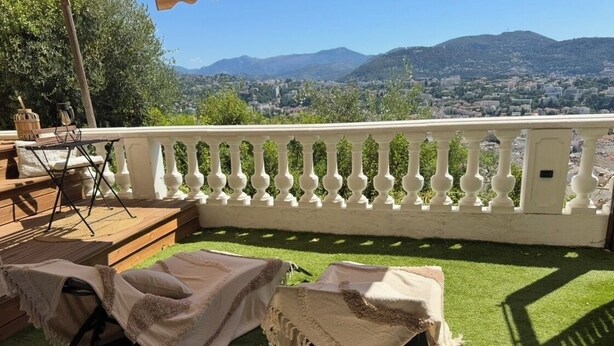 Eine Terrasse mit zwei Sitzmöglichkeiten, einem Tisch und einer Brüstung mit Blick auf eine Stadtlandschaft.