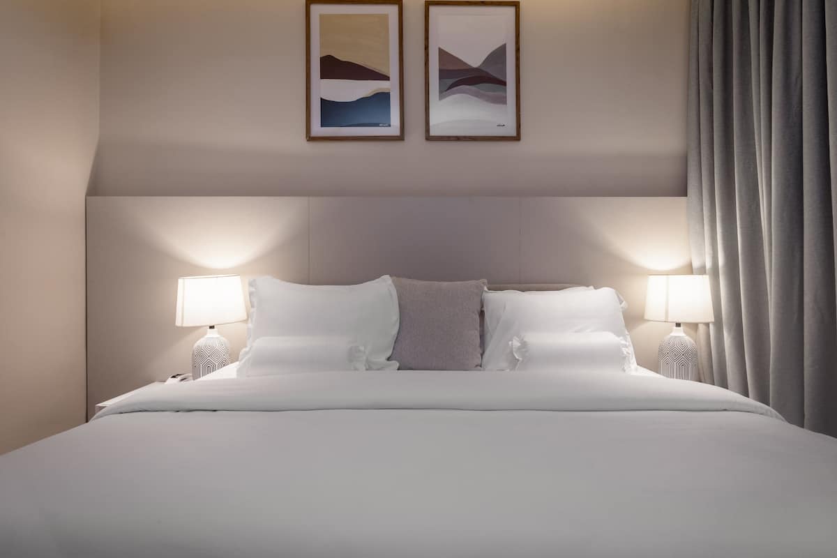 Un lit bien fait, avec du linge de lit blanc, deux lampes de chevet et deux tableaux abstraits encadrés accrochés au mur.