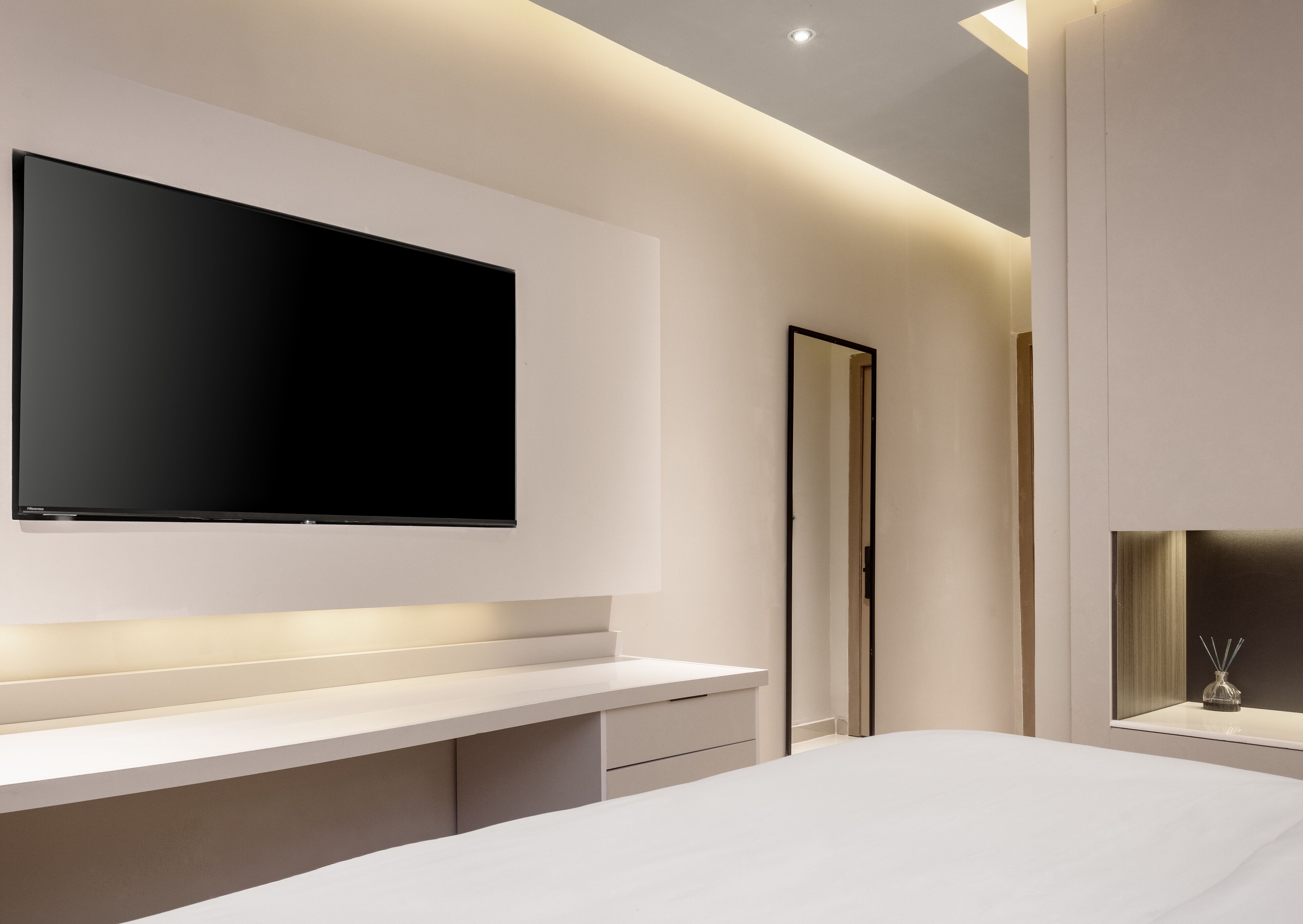 Un dormitorio moderno con una televisión de pantalla plana grande montada en la pared, una mesita de noche blanca y una cama con cabecera blanca.