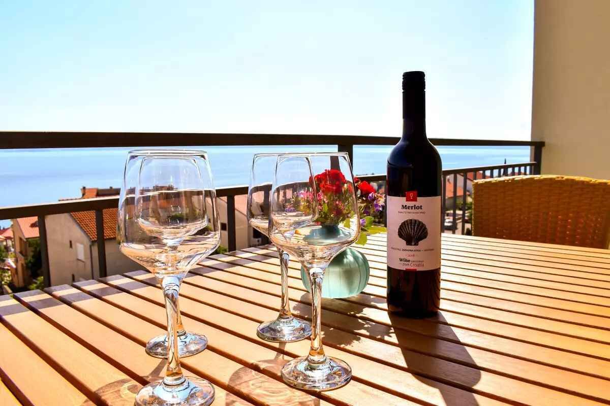 Een houten tafel met twee wijnglazen en een fles Merlot wijn op een balkon met uitzicht op zee.
