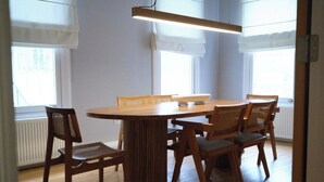 Ruokailutilassa on pyöreä puinen pöytä ja kuusi tuolia, suuret ikkunat, joissa on valkoiset verhot, sekä moderni riippuvalaisin.