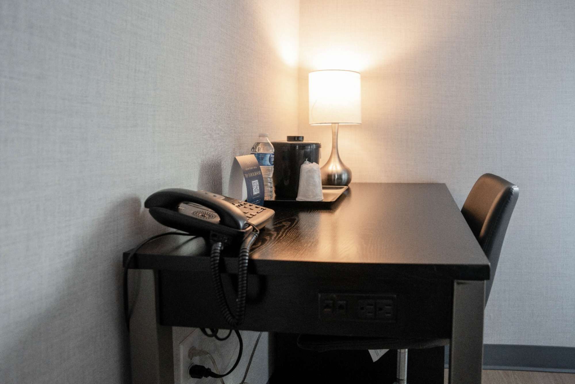 A hotel room with a desk, a phone, a lamp, a water bottle, and a coffee maker.