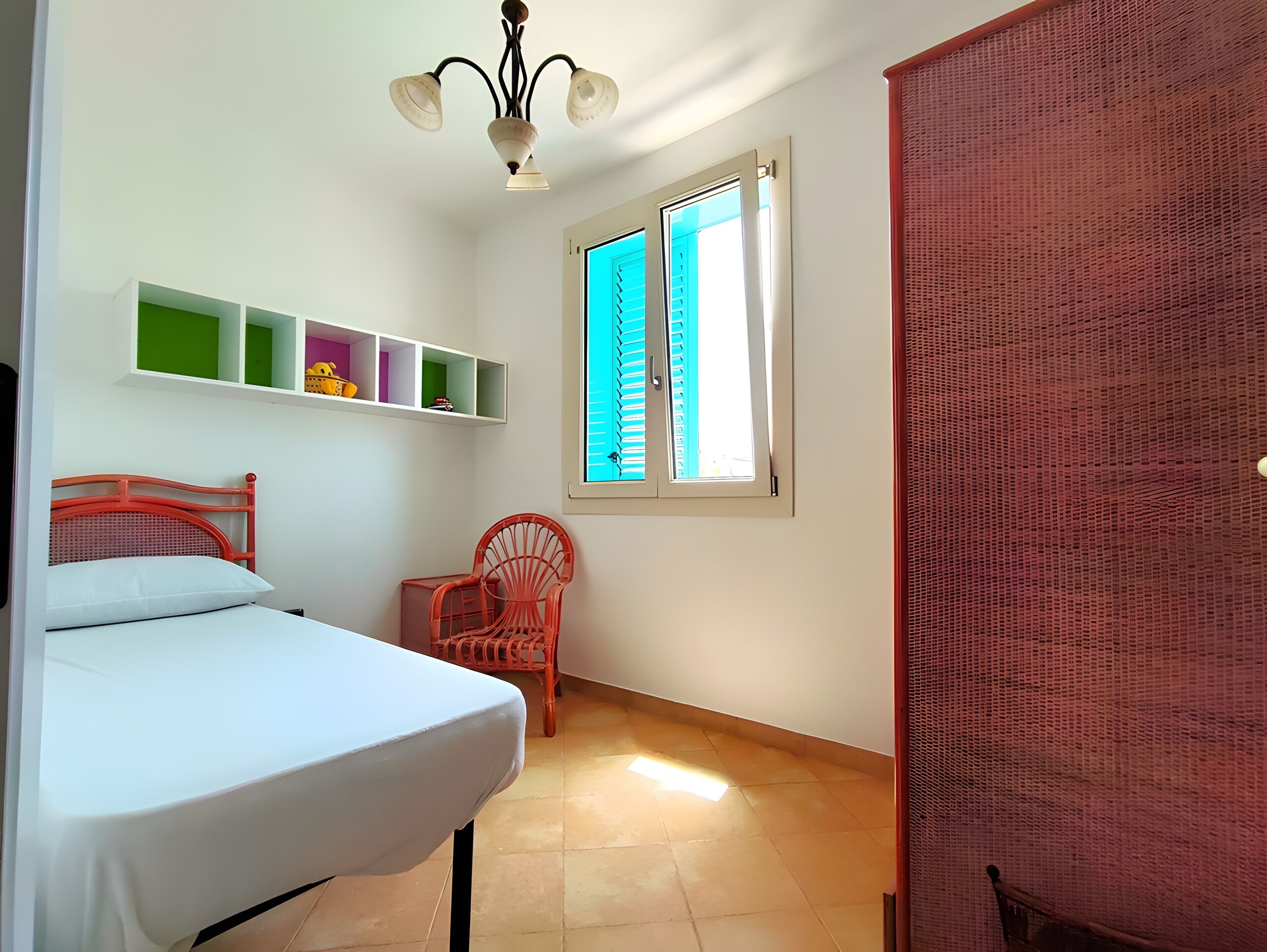 A room with a bed, a chair, a shelf with toys, a window with shutters, and a red door.