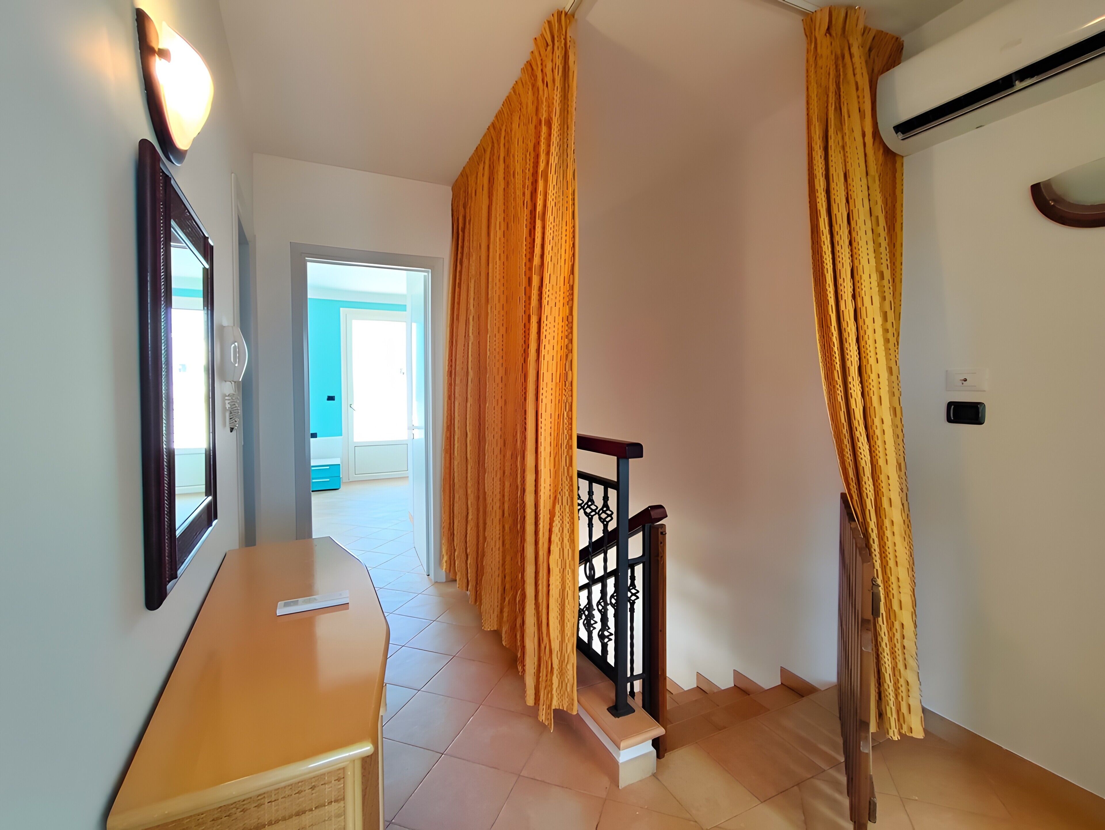 A hallway with a mirror, a light fixture, a staircase with a railing, and a door leading to another room.