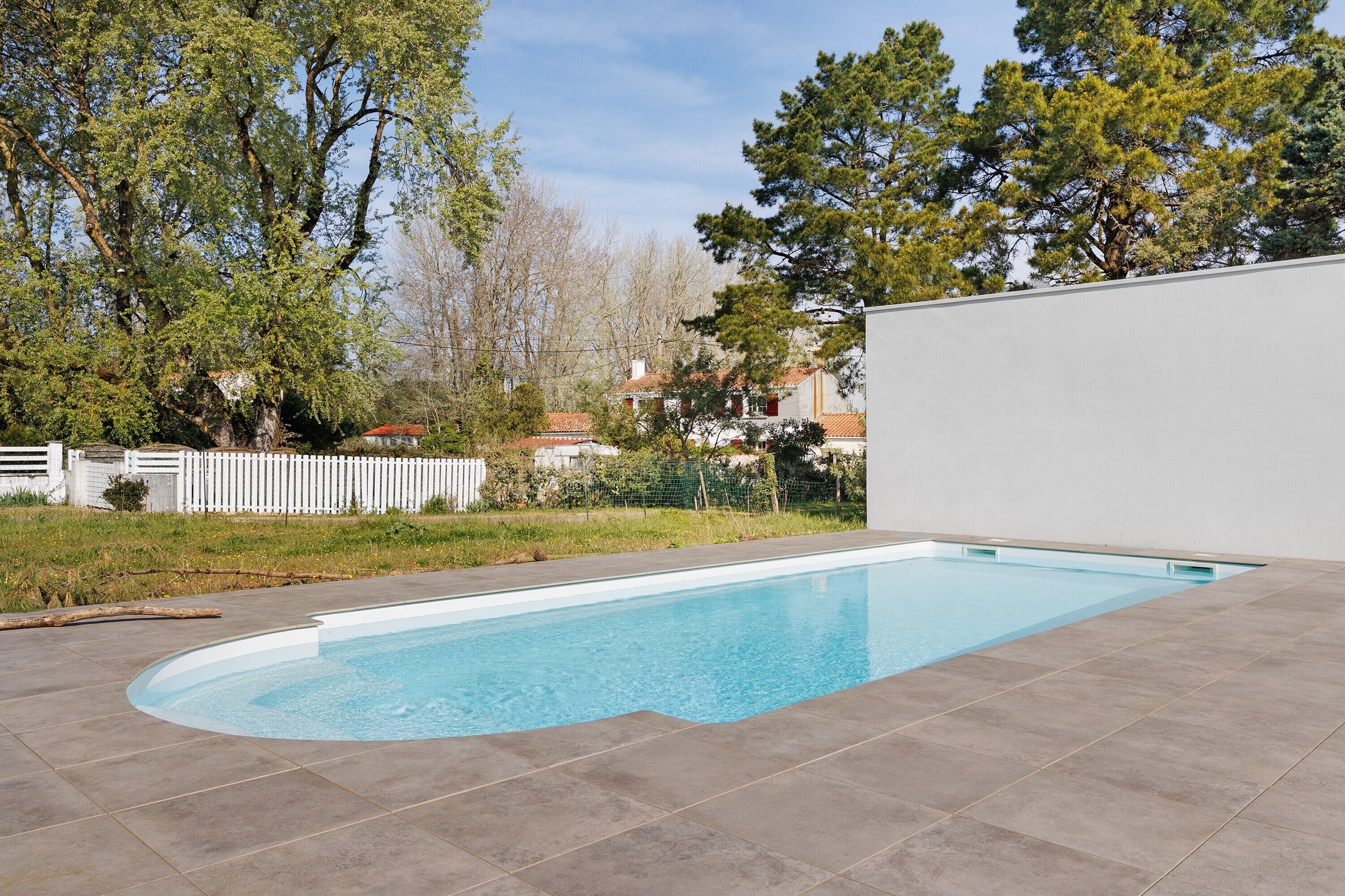 Una piscina rectangular con bordeado de azulejos, rodeada de árboles y una valla blanca.