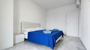 Un dormitorio con cama, mesita de noche, colcha azul y cabecera blanca.
