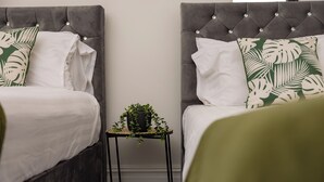 Dos camas con cabeceras grises, ropa de cama blanca y almohadas con estampado de hojas verdes. Una pequeña mesa negra con una planta está entre las camas.