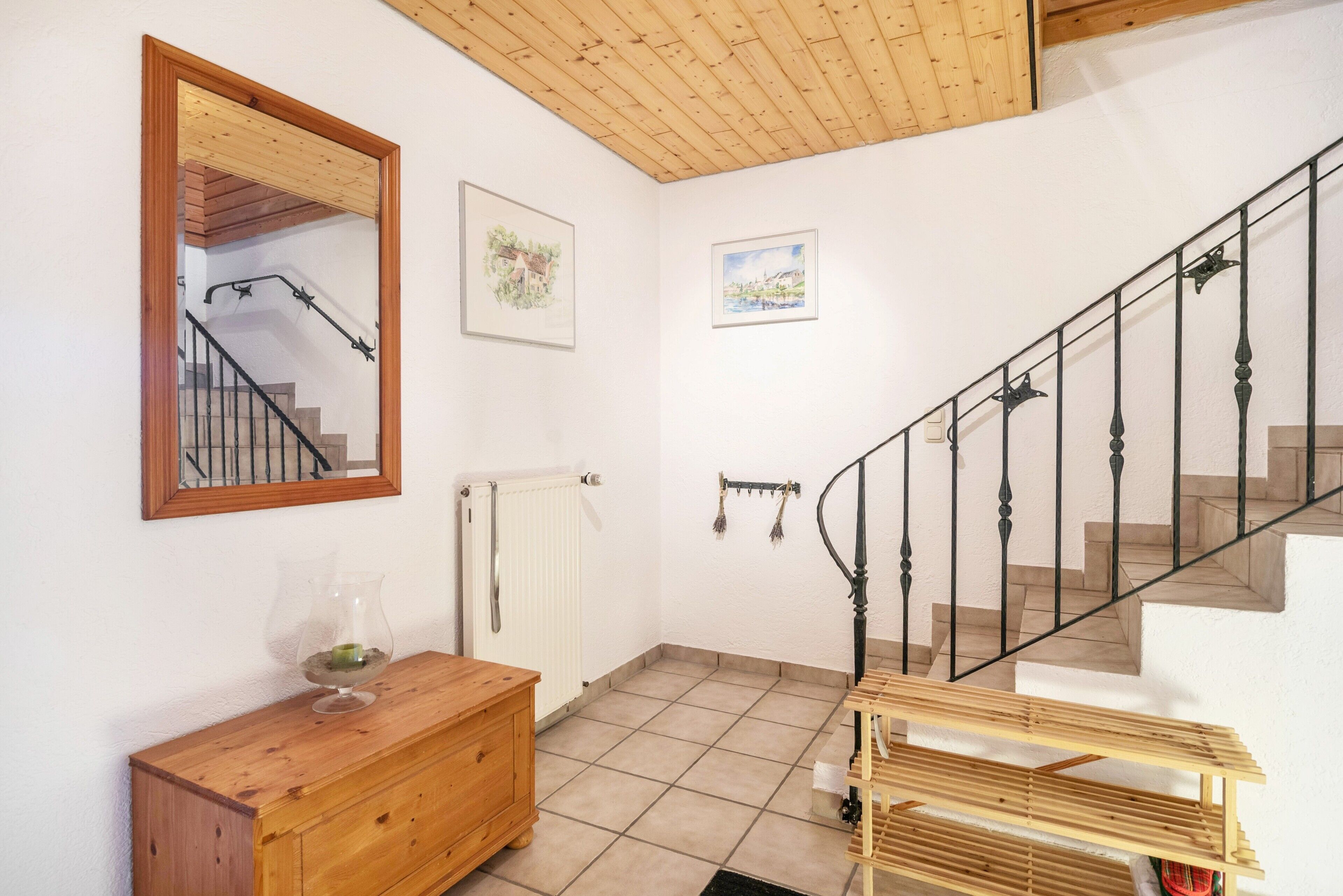 Una escalera con barandilla de madera y balaustres negros, un banco de madera, un armario de madera, un espejo y dos cuadros enmarcados en la pared.