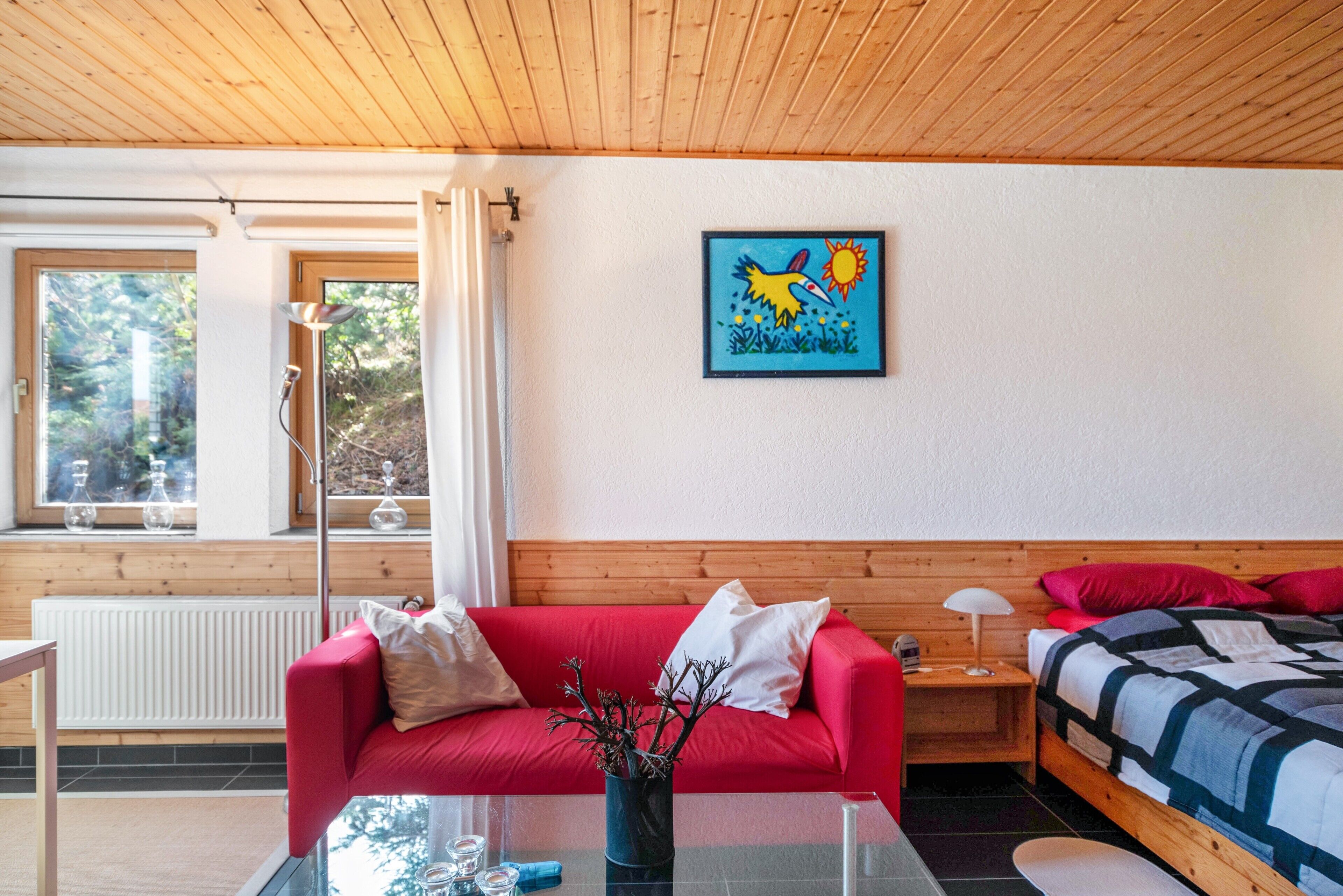 A room with a red sofa, a wooden headboard, a bed with a blue and white quilt, a painting of a bird, and a window with a view of trees.