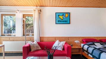 A room with a red sofa, a wooden headboard, a bed with a blue and white quilt, a painting of a bird, and a window with a view of trees.