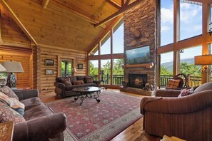 A log cabin interior with a fireplace, large windows, and a cozy seating area. - Among the Clouds | By Colonial Properties (Gatlinburg)