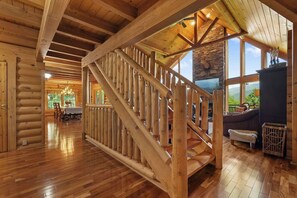A log cabin interior with a wooden staircase, dining area, and large windows. - Among the Clouds | By Colonial Properties (Gatlinburg)