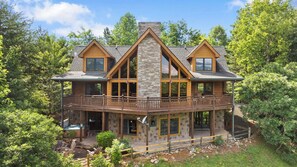 A two-story log cabin with a stone chimney, large windows, and a balcony. - Among the Clouds | By Colonial Properties (Gatlinburg)