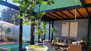 Un espacio interior moderno con un ventanal grande, una planta en maceta, una piscina y una zona de estar con mesa y sillas.