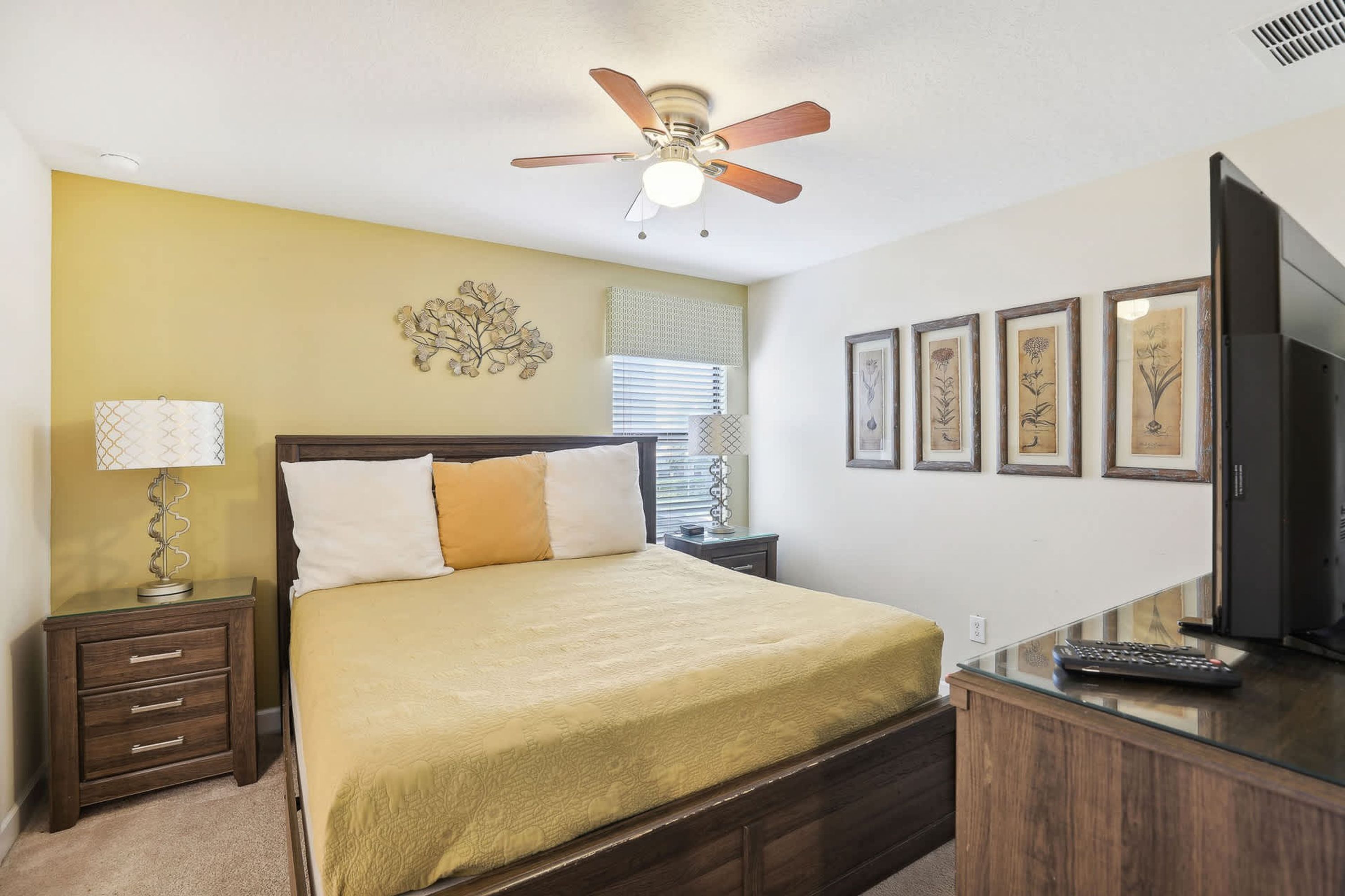 A bedroom with a bed, a nightstand, a desk, a ceiling fan, and framed botanical prints on the wall.