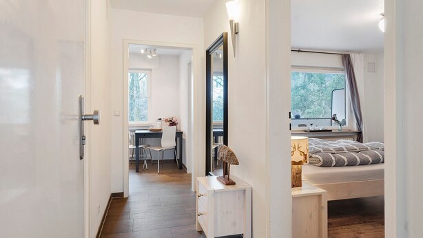 Een slaapkamer met een bed, nachtkastje en spiegel. Er is een bureau en stoel in de aangrenzende kamer.