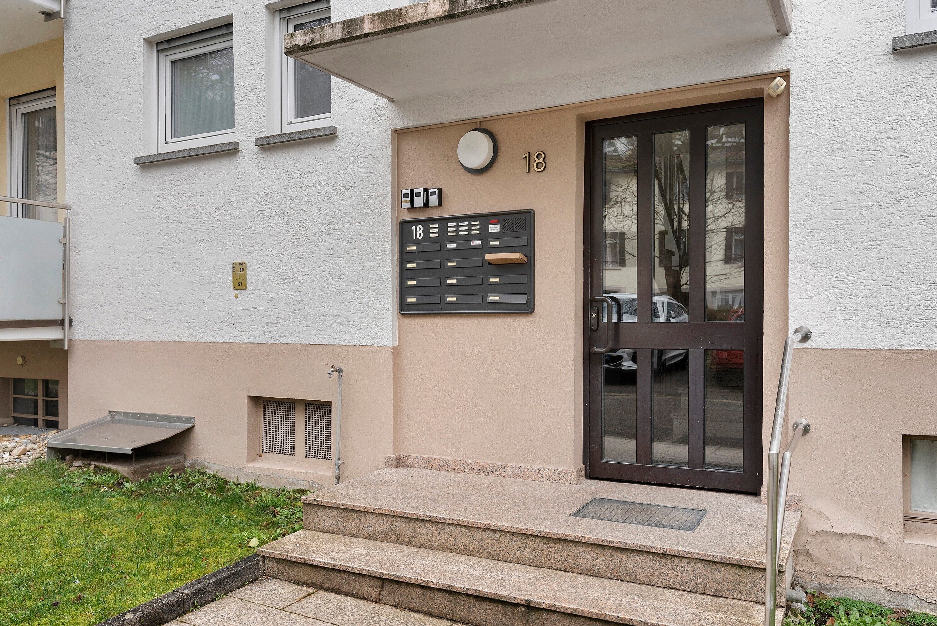 A building entrance with a numbered door, a small outdoor area, and a staircase leading up to the entrance.