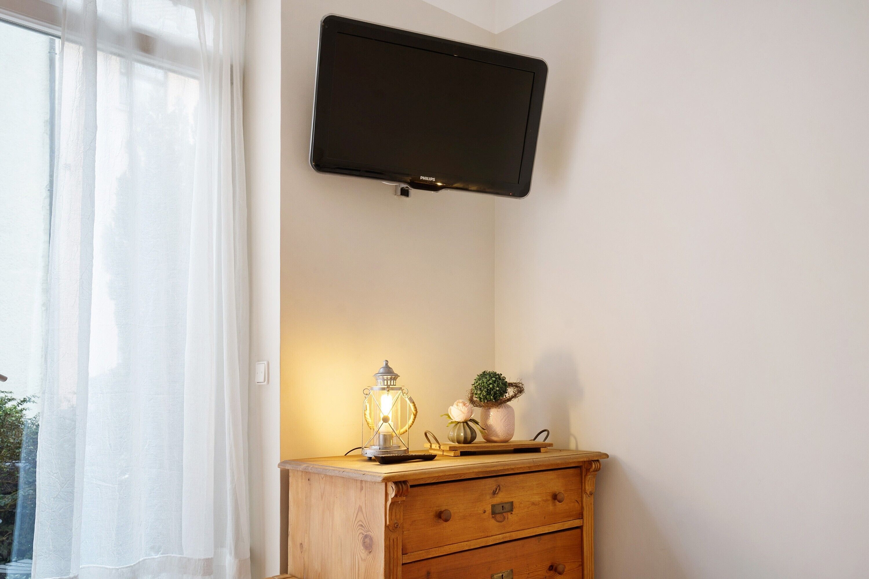 A room with a mounted television, a wooden dresser, a table lamp, and a potted plant.