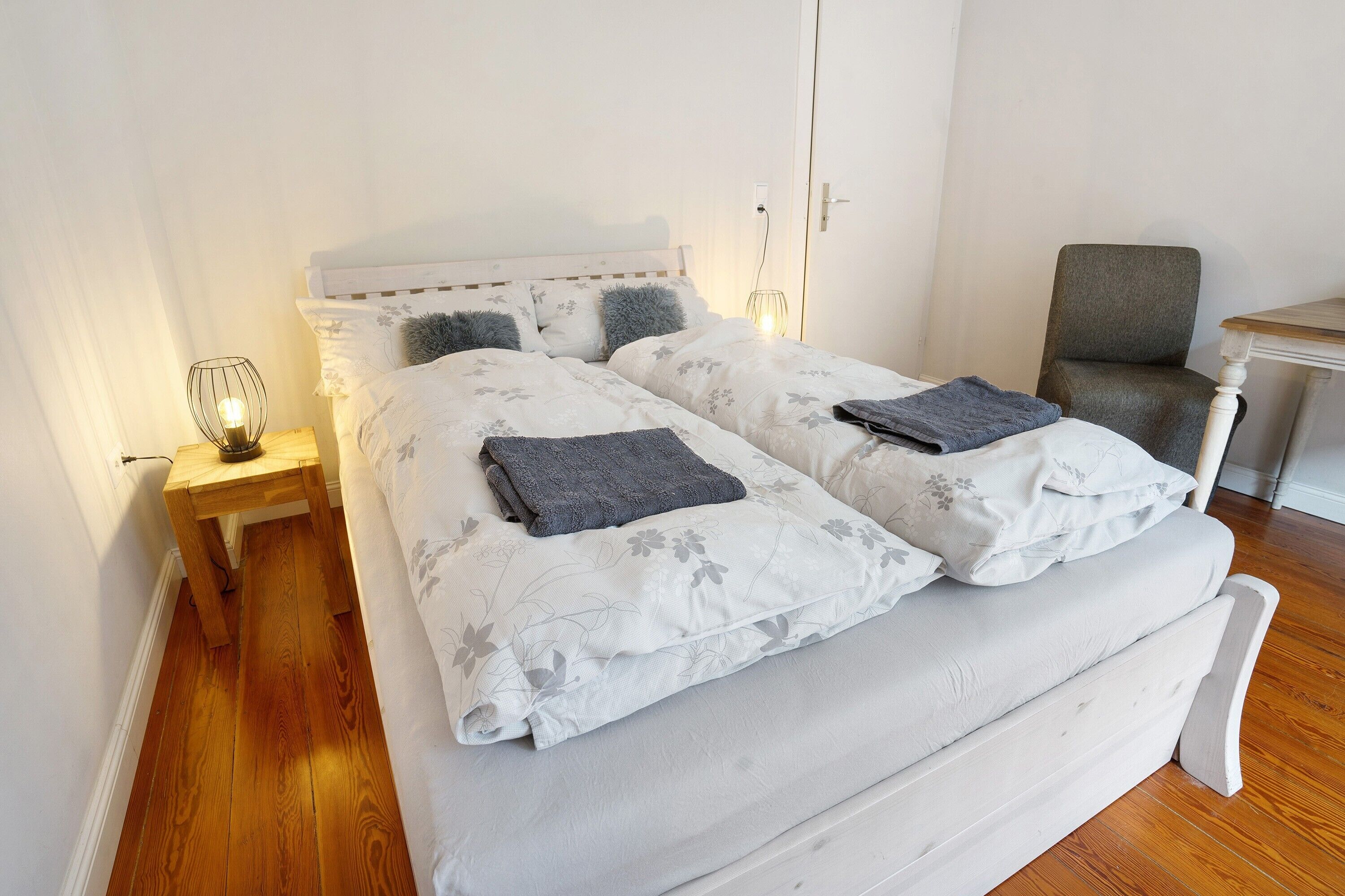 A double bed with floral bedding, a bedside table with a lamp, and a chair in a room with wooden flooring and white walls.