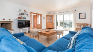 Un soggiorno con un grande divano blu, un tavolino in legno, una TV e un caminetto. È presente una porta scorrevole in vetro che dà su un balcone con mobili da esterno.