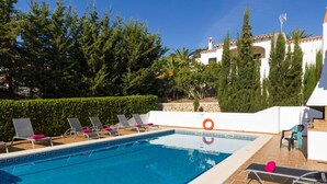 Una piscina rettangolare con superficie d'acqua blu trasparente, circondata da sdraio con cuscini rosa, un salvagente e un edificio bianco con tetto a tegole sullo sfondo.