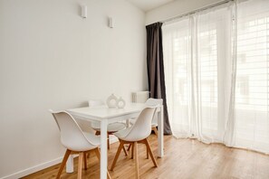 Habitació amb taula i cadires blanques, un radiador i una finestra amb cortines lleugeres.