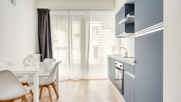 A modern kitchen with a white countertop, stainless steel appliances, and a window with sheer curtains. - Hostmate - Milano - Imperia's Pearl - Romolo IULM (Milano)