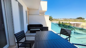 En balkon med et bord, stole, en sofa og udsigt til gaden og grønne områder.