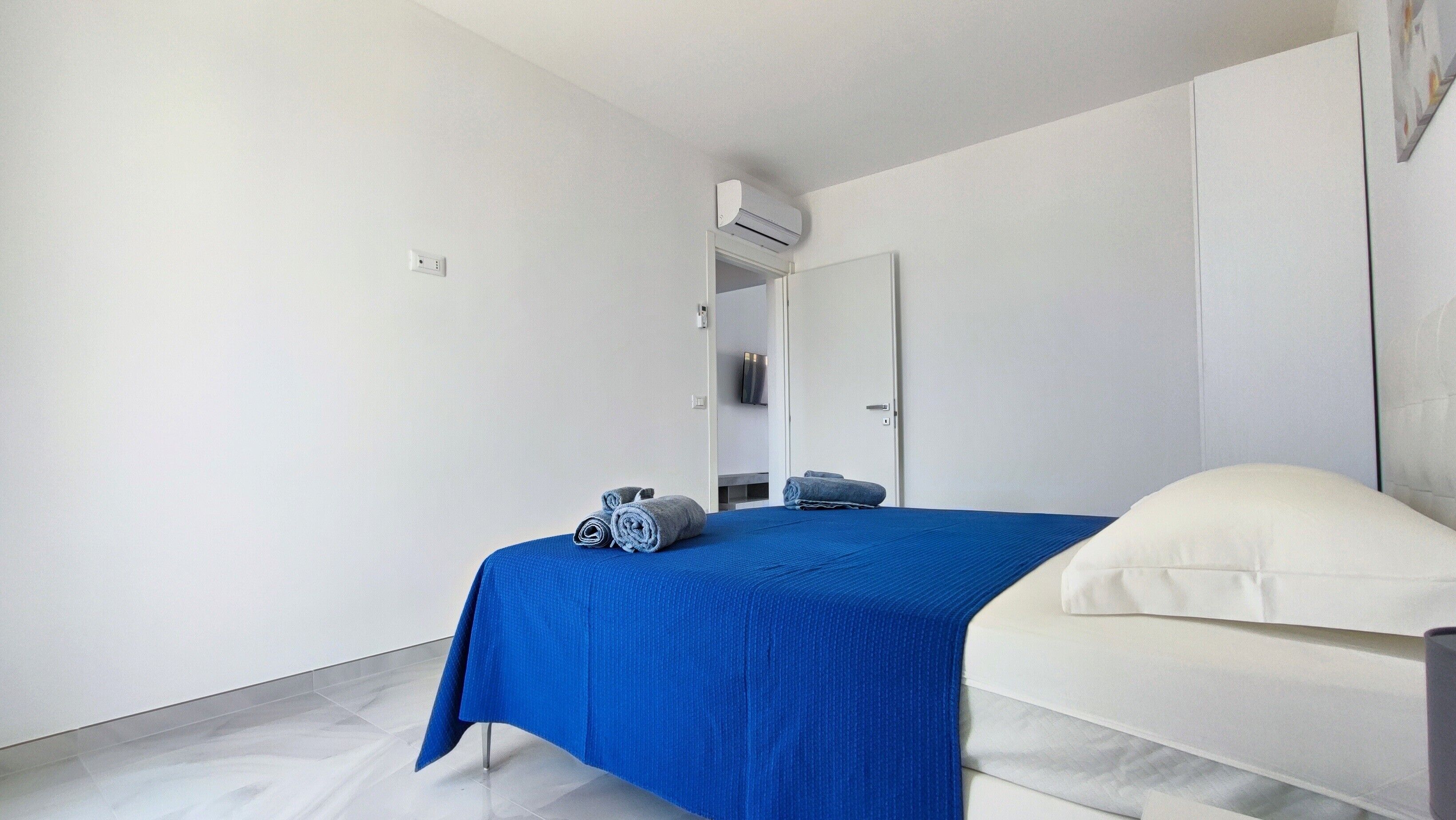 Un dormitorio moderno con una cama, una colcha azul, un almohadón blanco, una mesita de noche blanca y un equipo de aire acondicionado de pared.