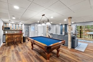 A pool table in a room with wooden flooring, a kitchen area with cabinets, and a view of the outdoors through large windows. - Lizard Creek Retreat | Pet-Friendly | New Boathouse (Littleton)