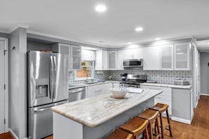 A modern kitchen with stainless steel appliances, white cabinetry, a marble countertop, and wooden stools. - Lizard Creek Retreat | Pet-Friendly | New Boathouse (Littleton)