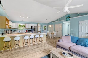 Eine moderne Küche mit weißen Schränken, einer zentralen Kochinsel mit Sitzgelegenheiten, einem Essbereich mit Tisch und Stühlen sowie einem Wohnzimmer mit Sofa und Deckenventilator.