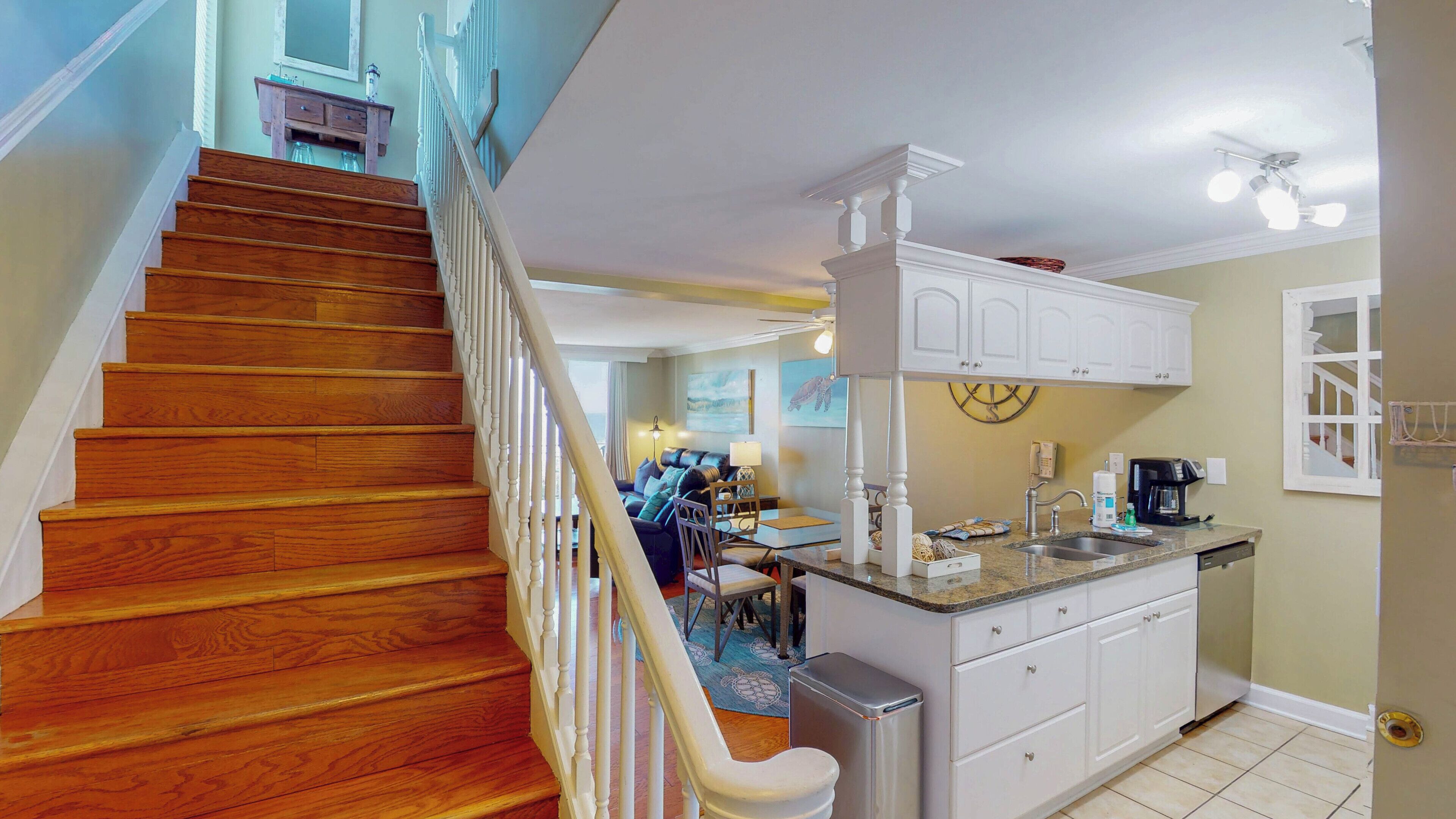A spacious kitchen with a wooden staircase, white cabinetry, and a dining area with a view of the living room.
