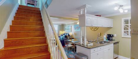 A spacious kitchen with a wooden staircase, white cabinetry, and a dining area with a view of the living room.