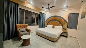 A modern bedroom with a large bed, a tufted armchair, a round coffee table, and a ceiling fan. - White Panda Hotels Shree Radhe (Dwarka)