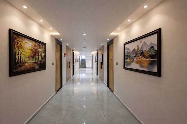 A hotel corridor with framed artwork on the walls, a polished floor, and a staircase at the end. - White Panda Hotels Shree Radhe (Dwarka)