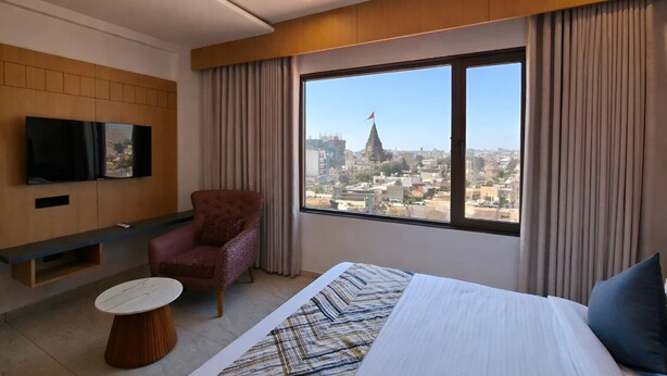 A hotel room with a bed, a chair, a TV mounted on the wall, and a large window offering a city view. - White Panda Hotels Shree Radhe (Dwarka)