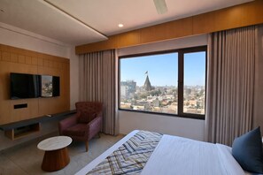 A hotel room with a bed, a chair, a TV mounted on the wall, and a large window offering a city view. - White Panda Hotels Shree Radhe (Dwarka)