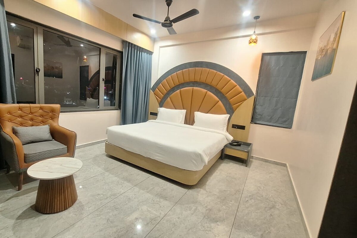 Un dormitorio moderno con una cama grande, un sillón capitonado, una mesita redonda y un amplio ventanal con cortinas.