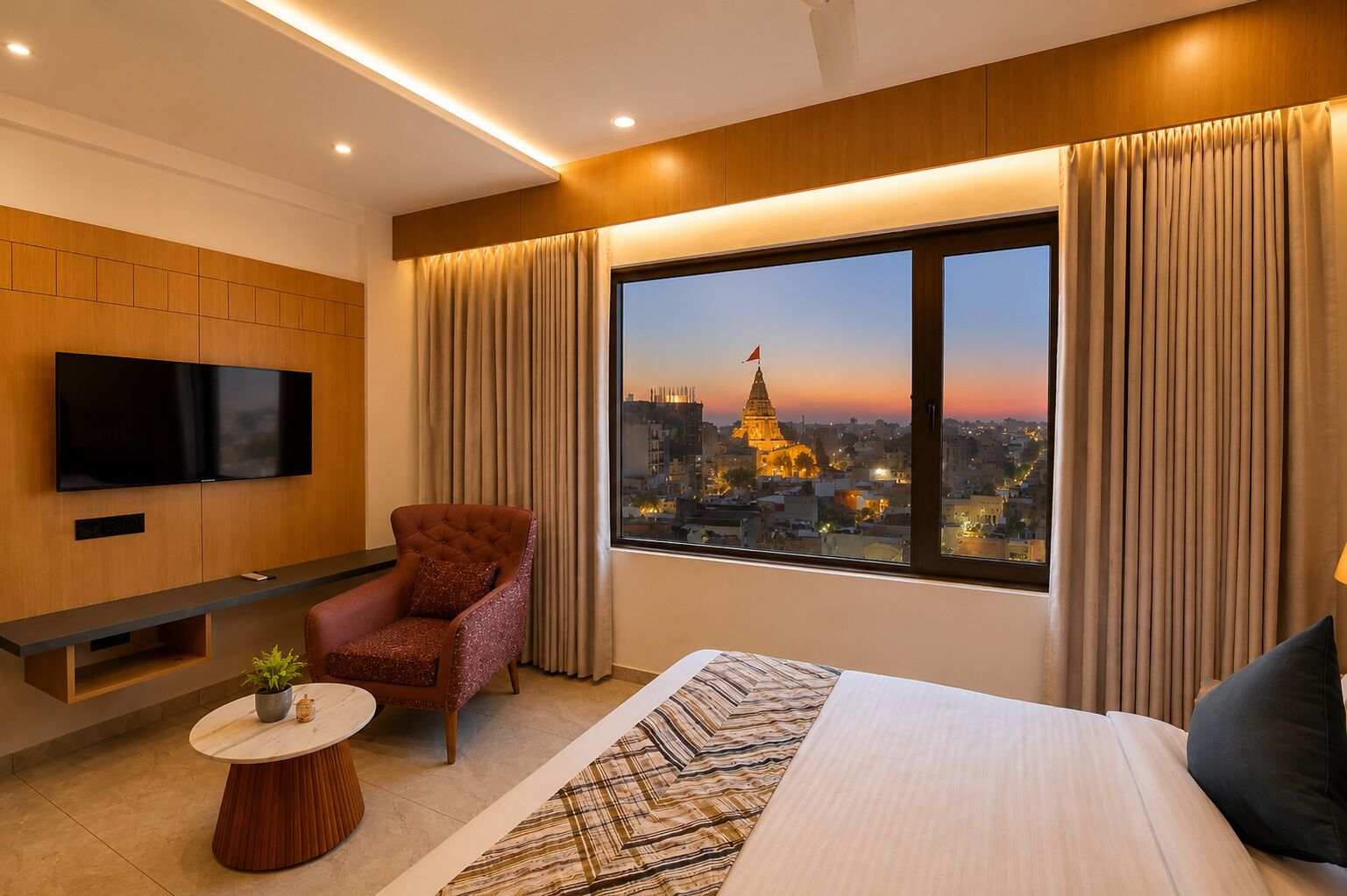 Une chambre d’hôtel moderne avec un grand lit, un téléviseur fixé au mur, un fauteuil confortable, une petite table ronde agrémentée d’une plante, et une vue sur le paysage urbain au coucher du soleil.