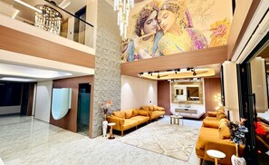 A spacious living area with a high ceiling, a large sofa set, a flat-screen TV, and a decorative wall mural. - White Panda Hotels Shree Radhe (Dwarka)