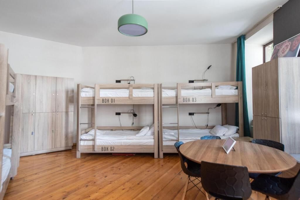 Ein Schlafsaalzimmer mit Etagenbetten, beschriftet mit BOX 01, BOX 02 und BOX 03, einem runden Holztisch mit Stühlen und einer grünen Pendelleuchte.