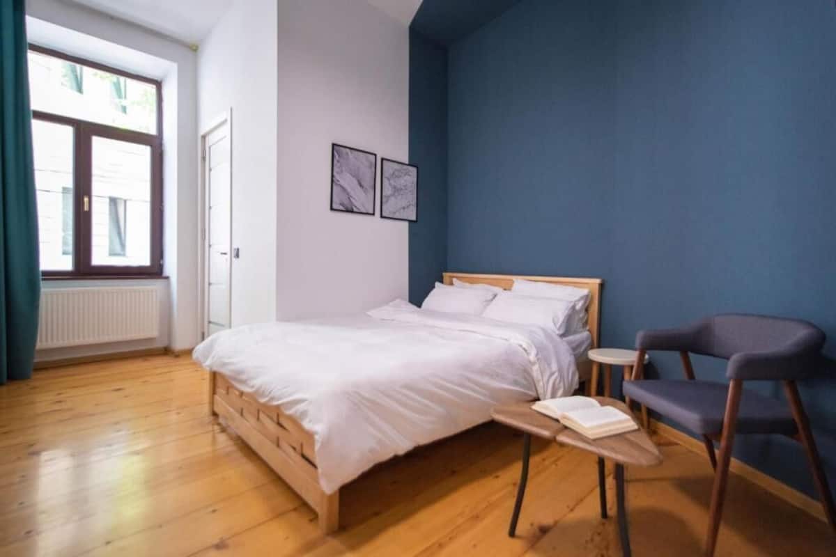 A bedroom with a wooden bed, a small bedside table with a book, a chair, and two framed pictures on the wall.