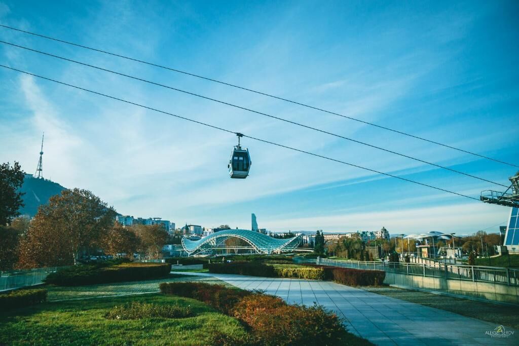 A cable car moving over a bridge in a park area with greenery and buildings in the background.