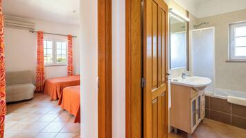 A hotel room with a wooden wardrobe, a bathroom with a sink and mirror, and two beds with orange bedding.