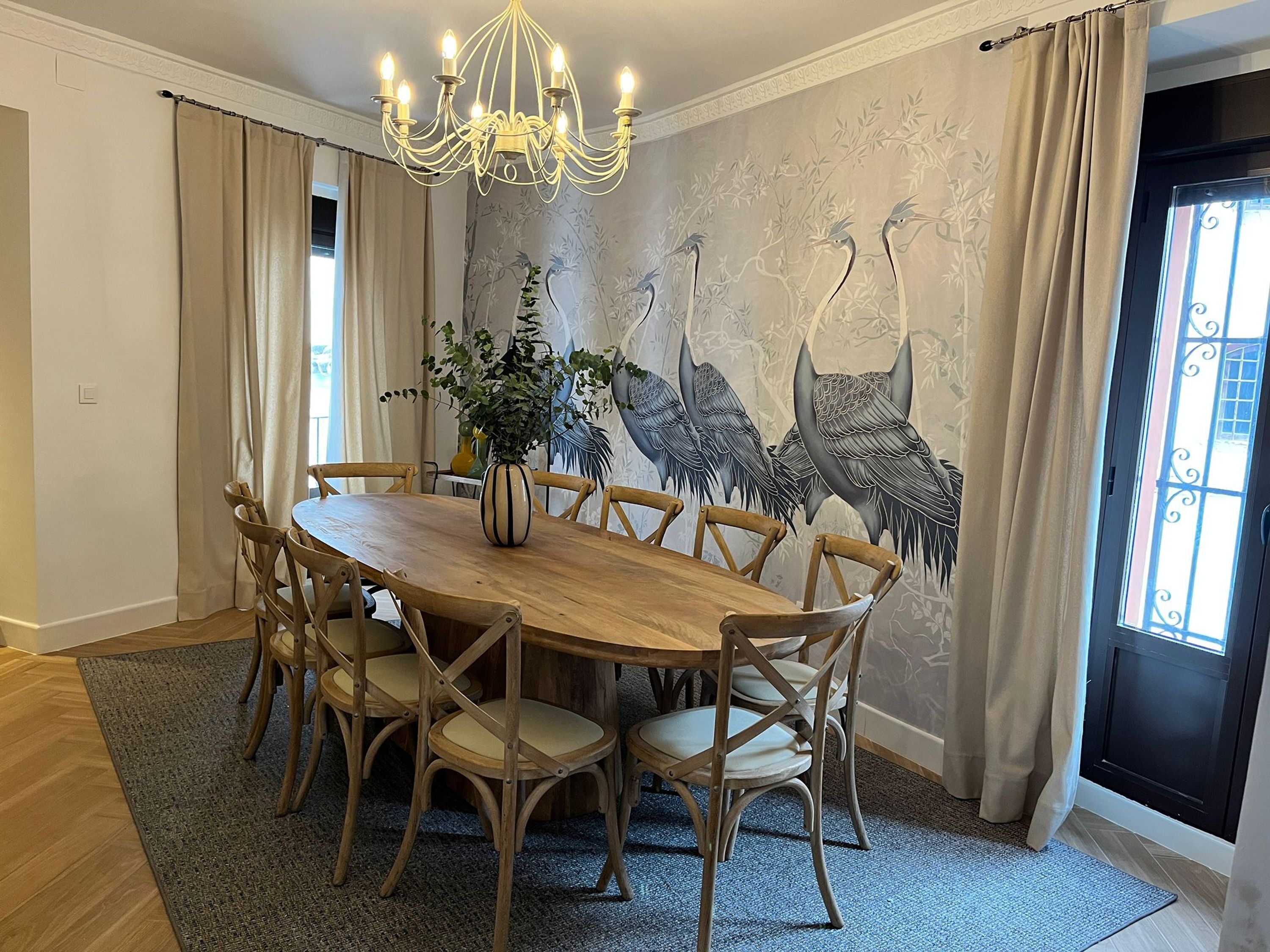 Un comedor con una mesa y sillas de madera, un candelabro, un mural de aves en la pared y una ventana con cortinas.
