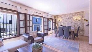 Un'ampia zona giorno con divano, tavolino da caffè e tavolo da pranzo con sedie. Grandi finestre con tende, una parete in pietra e vista esterna.