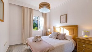 Un dormitorio ordenado con una cama grande, una mesita de noche con lámpara y una ventana con cortinas.