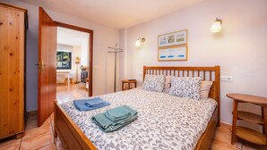 A bedroom with a bed, wooden headboard, two wall-mounted lights, a small table, and a door leading to another room.